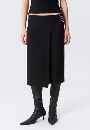 Jupe portefeuille noire mi-longue avec découpe latérale et double boucle, portée avec des bottes noires à bout pointu montant jusqu'au genou.