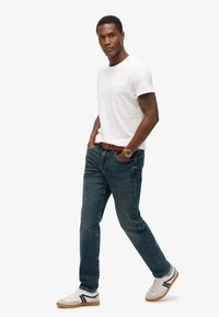 Camiseta blanca de manga corta, jeans de mezclilla azul, cinturón de cuero marrón, reloj de pulsera con esfera dorada y zapatillas blancas con detalles en negro.