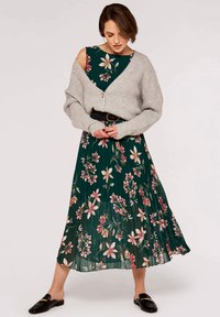 Blumenkleid mit einem grünen Grundton und rosa Blumen, kombiniert mit einer hellgrauen Rippstrickjacke und einem schwarzen Gürtel. Schwarze Slipper.