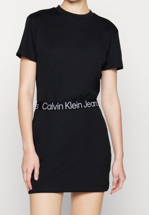 Kvinde iført en kortærmet sort kjole med elastik i taljen med "Calvin Klein Jeans" skrevet, stående mod en ensfarvet hvid baggrund.