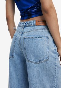 Light blue denim jeans with a relaxed fit, featuring two back pockets, a brown leather logo patch, and subtle texture variations.