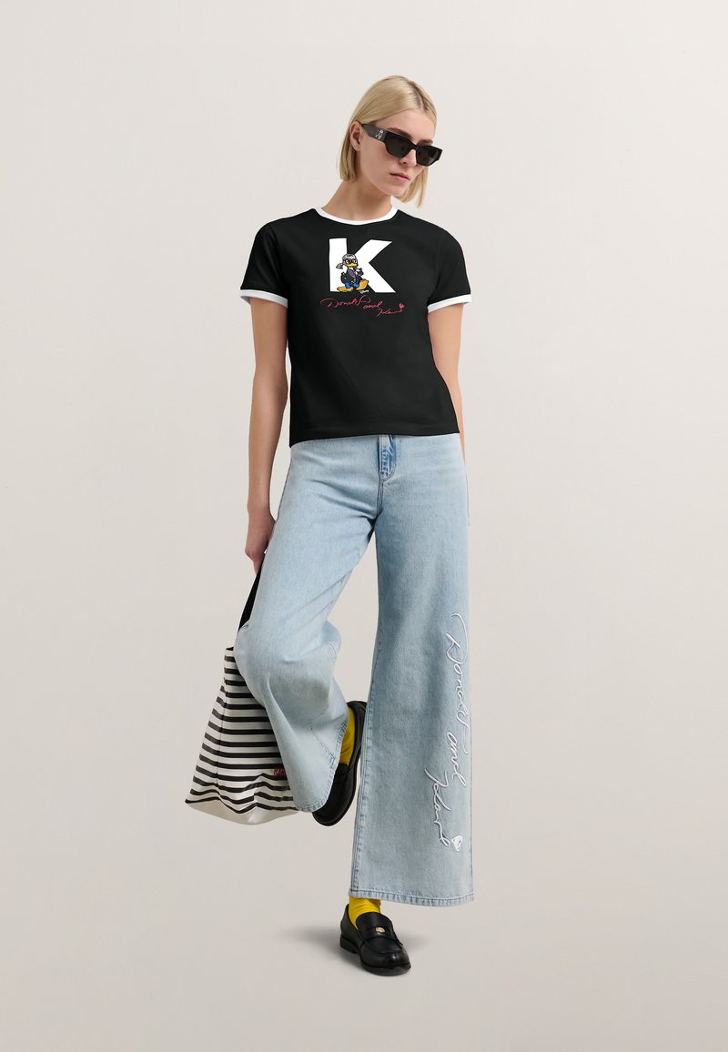 Czarna koszulka z białymi wykończeniami, z dużą literą "K" i rysunkowym wzorem, w zestawie z szerokimi jasnoniebieskimi dżinsami. Białe hafty na dżinsach.