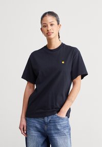 Sort bomulds t-shirt med rund hals og korte ærmer. Har et lille gult logo på brystet. Kombineret med lyseblå jeans.