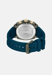 Orologio con cinturino in gomma blu e retro in acciaio inossidabile, quadrante argento con la scritta "POLICE", dettagli dorati e design testurizzato sul retro.