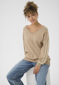 Beiger V-Ausschnitt-Pullover mit langen Ärmeln, der cremefarbene Streifen entlang der Ärmel aufweist; getragen mit blauen Jeans. Glatte Textur, lässiges Design.