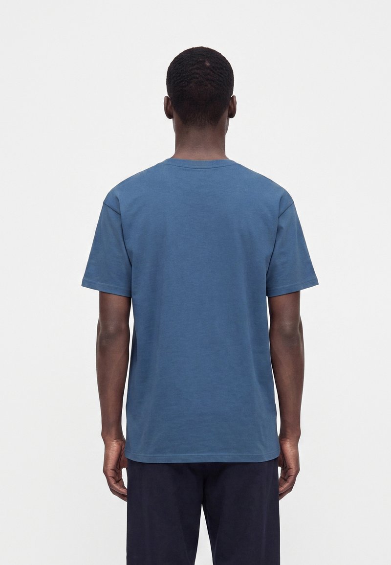 Mann von hinten gesehen, trägt ein schlichtes blaues T-Shirt mit kurzen Ärmeln und dunkle Hose vor einem neutral grauen Hintergrund.