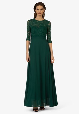 Lange groene jurk met een kant bodice met een bloemenpatroon en driekwart mouwen, met een vloeiende chiffon rok en platte zwarte sandalen.