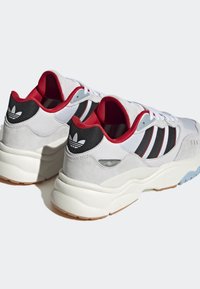 Ett par vita och grå Adidas-sneakers med svarta ränder, röd inredning och tjocka vita sulor på en ljus bakgrund.