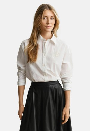 Femme aux longs cheveux blonds portant une chemise blanche boutonnée et une jupe en cuir noire, debout devant un fond clair uni.