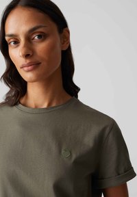 T-shirt en coton vert olive avec un col rond, des manches roulées et un petit accent de cœur brodé dans une teinte plus foncée de vert.