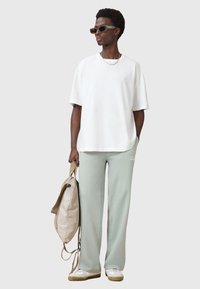 Hvid oversized t-shirt, lysegrøn sweatpants med logo, solbriller og beige rygsæk. Glat stof, afslappet pasform, minimalistisk design.