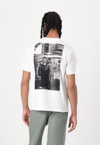 White cotton T-shirt featuring a large black-and-white photographic print of people at a bus stop on the back. Crew neckline, relaxed fit.