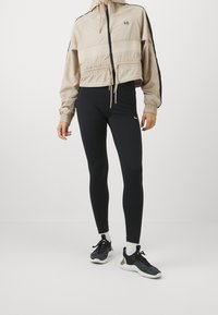 Lys beige cropped jakke med elastisk talje, sorte detaljer og hætte. Sorte leggings med glat tekstur og subtilt Nike-logo. Sorte sneakers.