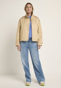 Beige cropped jacket with a high collar, button closures, and side pockets, paired with light blue loose jeans and beige sneakers.