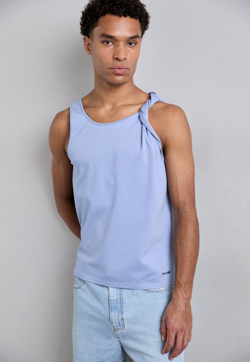 Lichtblauw, mouwloos tanktop met een geknoopt detail op een schouder, gemaakt van een gladde stof. Gecombineerd met lichtblauwe denim shorts.