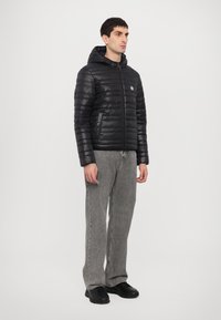 Chaqueta de plumas negra con capucha, textura acolchada, cremallera delantera, acento de logo en el pecho, combinada con pantalones grises de corte holgado y zapatos negros.