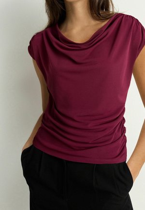 Kobieta w bezrękawowej fioletowej bluzce z drapowanym dekoltem i czarnych spodniach, z rękami w kieszeniach, stojąca na jednolitym tle.