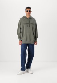 Felpa verde oliva con tasca frontale, logo "TOMMY JEANS", abbinata a jeans blu e sneakers bianche. Il modello indossa occhiali da sole rettangolari.