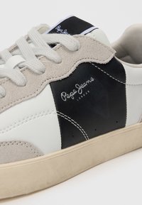 Sneaker in pelle bianca e nera con dettagli in camoscio grigio, lacci piatti e suola in gomma beige. Presenta una cucitura con il logo sul lato.