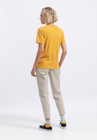 Geel katoenen T-shirt, korte mouwen, met een klein logo-detail op de achterkant. Gecombineerd met beige broeken en zwarte sneakers met kleurrijke sokken.