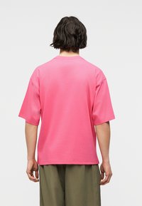 Άτομο με κοντά σκούρα μαλλιά, φορώντας ροζ κοντομάνικη μπλούζα και ελιάς πράσινο παντελόνι, εμφανιζόμενο από πίσω.