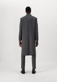Cappotto grigio con vestibilità sagomata, lungo, a un petto e con spalle strutturate. Indossato sopra pantaloni grigi abbinati e stivali.