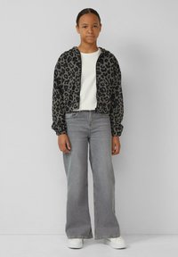 Korte jas met zwart en grijs luipaardprint, gebreid materiaal, elastische manchetten; gecombineerd met grijze wijde jeans en witte sneakers.