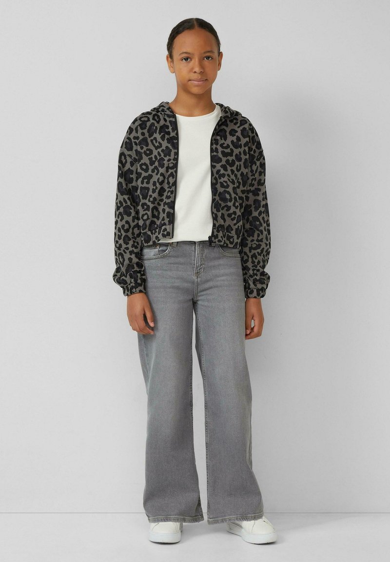 Korte jas met zwart en grijs luipaardprint, gebreid materiaal, elastische manchetten; gecombineerd met grijze wijde jeans en witte sneakers.