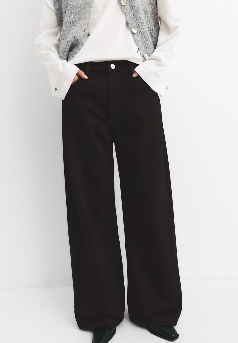 Personne portant un pantalon noir à jambes larges, une chemise blanche à manches longues et un gilet en tricot gris boutonné, les mains dans les poches.