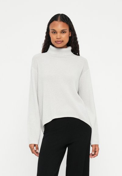 Pull à col roulé gris clair avec une texture côtelée, coupe oversize et manches longues ; associé à un pantalon noir à jambes larges.