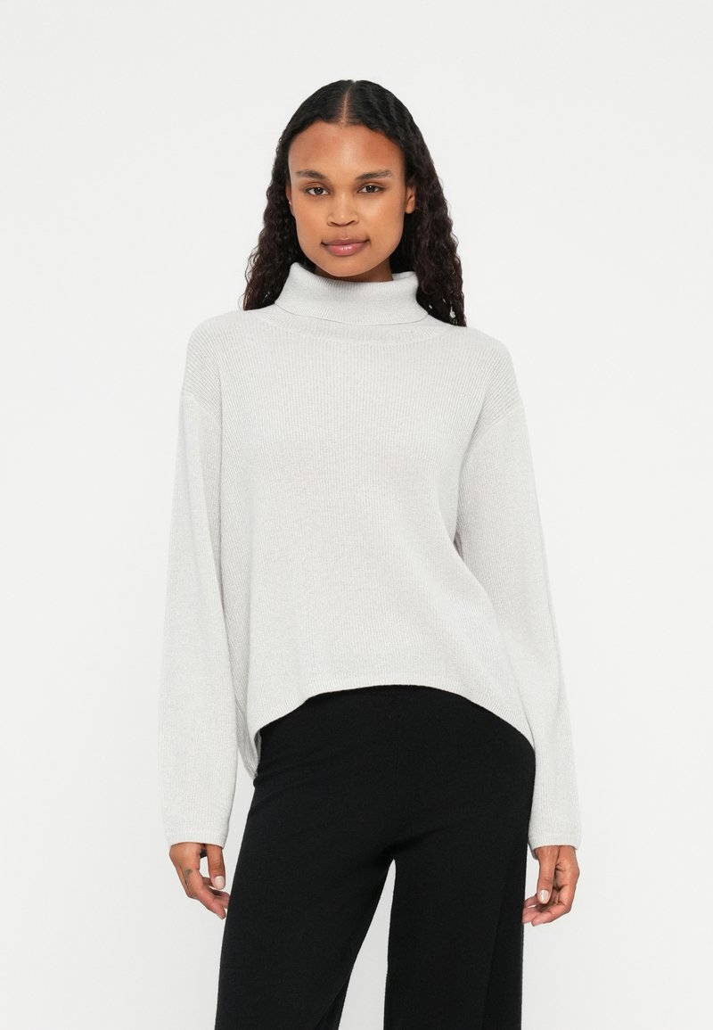 Pullover cinza-claro de gola alta com textura canelada, ajuste oversized e mangas longas; combinado com calças pretas de perna larga.