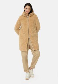 Fuzzy beige gevoerd jack met een rits, voorzakken en een gestructureerde buitenkant, gecombineerd met beige broeken en lichte sneakers.