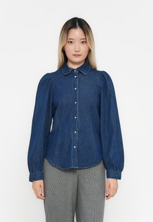 Denim shirt with a button-down collar, long puff sleeves, and a rounded hem. The fabric is dark blue with a smooth texture and subtle stitching.
