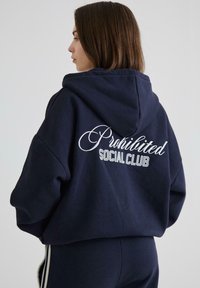 Marineblå hættebluse med "Prohibited Social Club" broderet i hvid og sølv på ryggen. Har en afslappet pasform og blød tekstur.