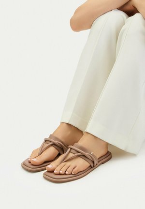 Voeten met beige platte sandalen met gekruiste bandjes en een witte broek tegen een effen lichte achtergrond.