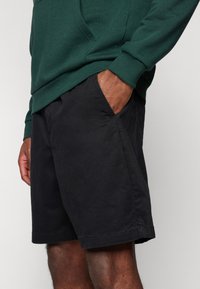Sudadera verde con bolsillo tipo canguro y puños acanalados, combinada con pantalones cortos negros que tienen bolsillos laterales y un ajuste relajado.