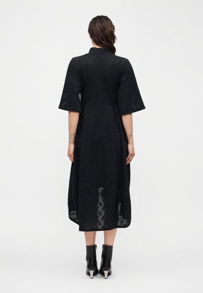 Γυναίκα με σκούρα μαλλιά φοράει μαύρο μίντι φόρεμα με μανίκια μέχρι τον αγκώνα και υφασμάτινο σχέδιο, συνδυασμένο με μαύρα μποτάκια αστραγάλου με τακούνια.