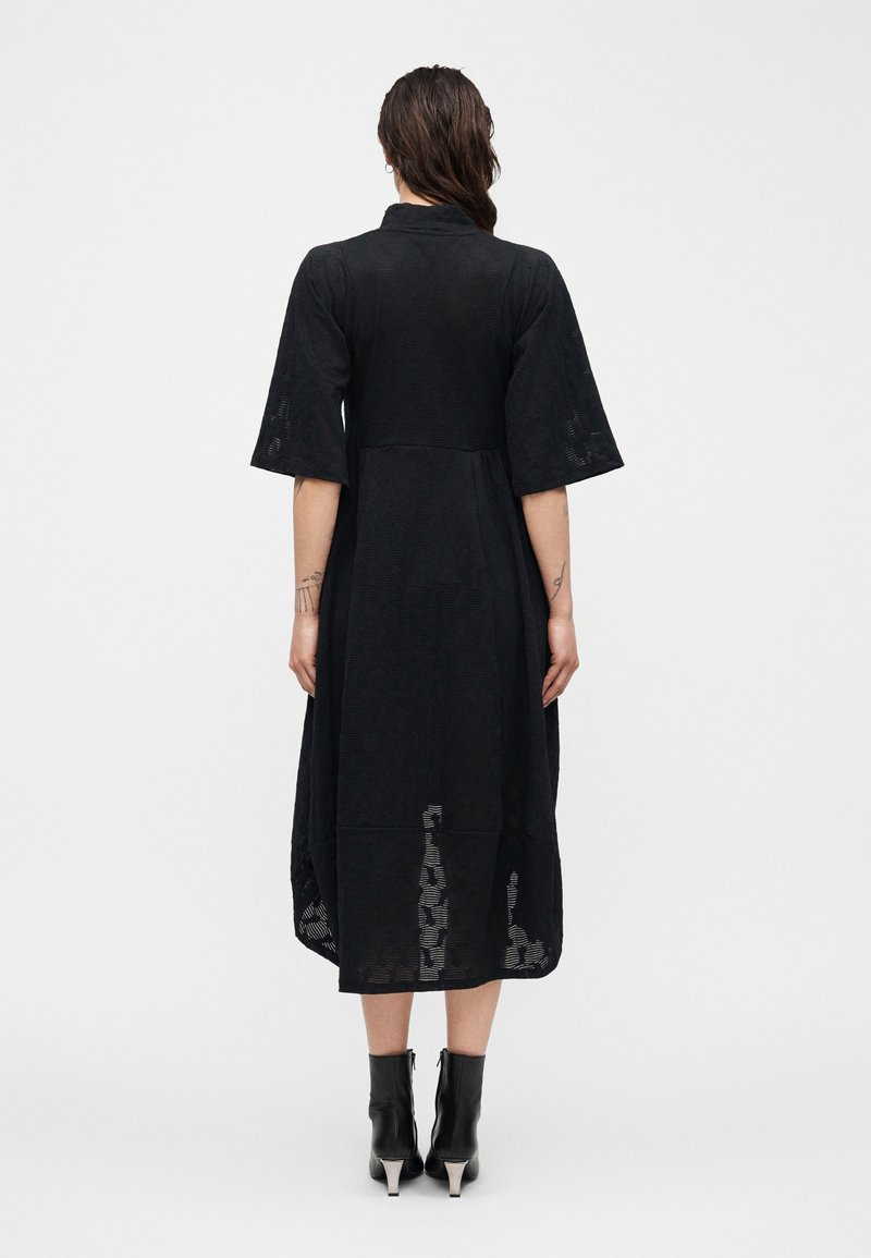 Frau mit dunklem Haar trägt ein schwarzes Midikleid mit Ellbogenlangen Ärmeln und gemustertem Stoff, kombiniert mit schwarzen Stiefeletten mit Absatz.