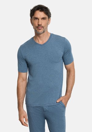 Mann mit kurzen dunklen Haaren und Bart, der ein blaues kurzärmeliges V-Ausschnitt-Shirt und passende Hose trägt, steht mit einer Hand in der Tasche.