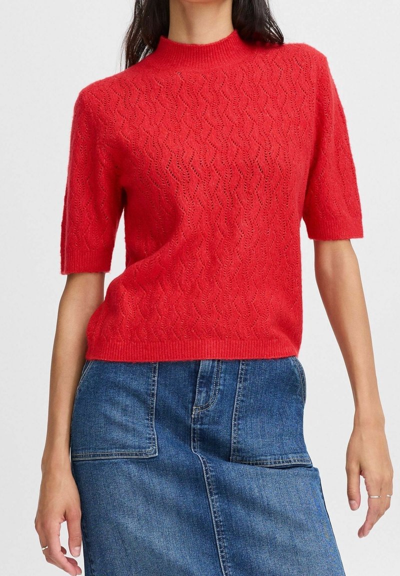 Femme portant un pull en maille rouge à manches courtes avec un motif texturé et une jupe en jean avec de grandes poches avant.