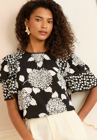 Blusa nera con motivo floreale bianco, maniche corte arricciate e orlo scallop. Caratterizzata da una vestibilità comoda e un tessuto leggero.