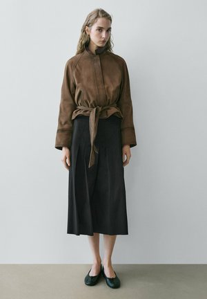 Femme aux cheveux clairs ondulés portant une veste ceinturée en suède marron, un pantalon large court noir et des ballerines noires, debout contre un mur uni.