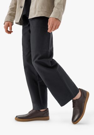 Scarpe di pelle marrone con finitura liscia, punta rotonda e suola in gomma marrone. Indossate con pantaloni scuri e larghi e una giacca beige.