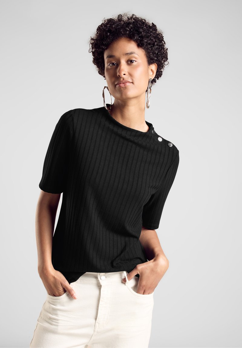 Haut noir côtelé à manches courtes avec un col montant et des boutons décoratifs sur l'épaule, associé à un pantalon de couleur claire.