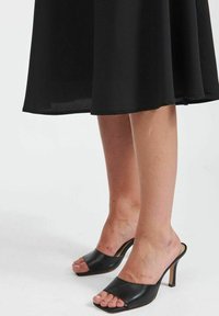 Mules ouvertes en cuir noir avec une large sangle sur le pied, dotées d'un talon haut effilé et d'une texture lisse.