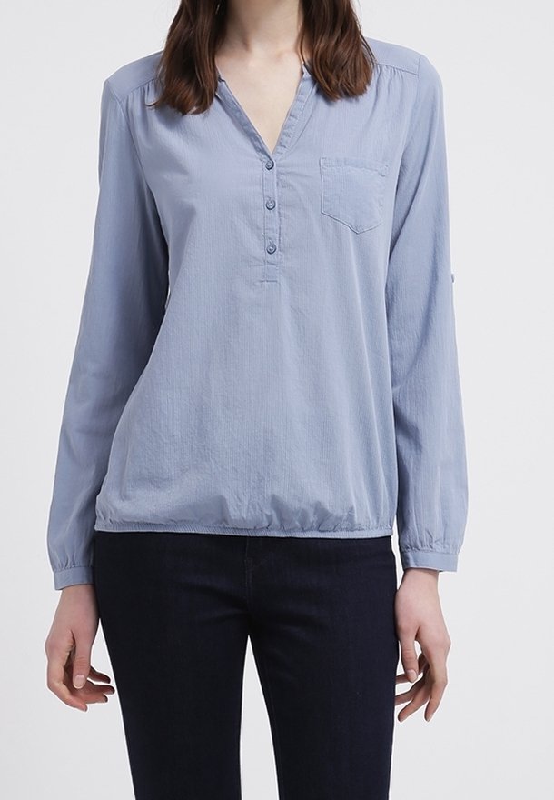 Blouse bleu clair à manches longues avec un col en V, fermeture boutonnée sur le devant et une petite poche poitrine; comprend un ourlet froncé et un tissu doux et texturé.