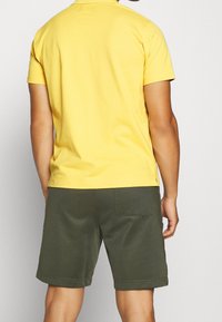 Homme portant une chemise à manches courtes jaune et un short vert olive, se tenant de dos face à un fond uni.