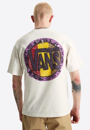Persona con capelli biondi corti indossa una t-shirt bianca Vans con un grande logo circolare multicolore sulla schiena e ha tatuaggi su entrambe le braccia.