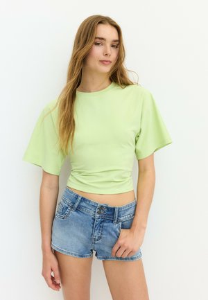 Ung kvinde med langt hår iført en lysegrøn tætsiddende top med korte ærmer og blå denimshorts, der står mod en hvid baggrund.