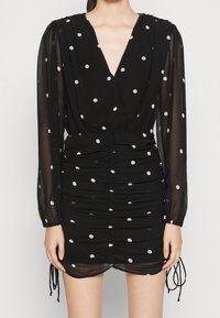 Svart klänning med djup V-ringning, långa genomskinliga ärmar, rynkade sidor och vita prickar. Inkluderar dragsko vid fållen.